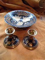 Painted brass bowl with floral pattern and pair of matching candle holders placed next to each other on brown fabric.