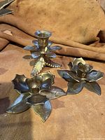 Three metal leaf-shaped candle holders displayed on a tan fabric background, showing their front profile and design.