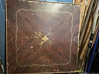 Top surface of square wooden card table showing ornate gold inlay and wood grain pattern with signs of wear.