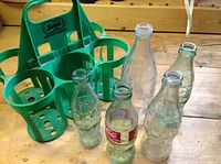 Five Coca-Cola glass bottles arranged next to the green plastic carrying case on a wooden surface.