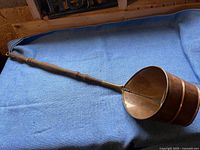 Full view of antique copper scooper with long wooden handle laid out on blue fabric.