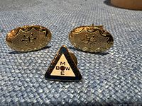 Front view of two gold-tone cufflinks and triangular MBOWE tie pin on blue fabric.