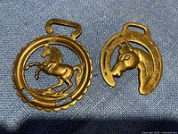 Three solid brass decorative horse brasses including one with a rearing horse inside a circular frame, one with a horse head and horseshoe, and one shaped as a horseshoe.