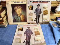 Front covers of Dolly Parton's "Coat of Many Colors" and Stompin' Tom Connors' "My Stompin' Grounds" vinyl LP albums on a blue surface.