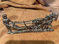 Photo of solid brass figurine showing a farmer guiding a horse pulling a plow, on a brown cloth background.