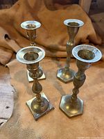 Front view of four brass candle holders of varying shapes and bases on light brown surface.