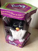 Furby in original hexagonal box, visible front portion with black and white plush, green eyes, and yellow beak.