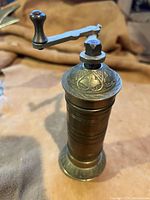 Front angle view showing detailed decorative engravings on lid and handle of the brass pepper grinder.