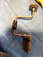 Full view of the antique hand drill showing metal crank arm with wooden handle and metal drill chuck on a blue cloth background.