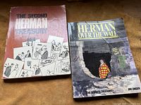 Two comic books, 'The Second Herman Treasury' and 'Herman Over the Wall' by Jim Unger, displayed side by side on a brown surface.