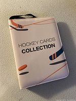 Front cover of the white hockey cards collection binder with hockey stick and puck graphics and text.