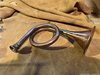 Side angle of the copper and brass horn on a tan cloth background, showing coiled circular shape and mouthpiece.