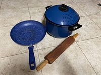 Photo showing three kitchenware items on floor: a large blue aluminum pot with matching lid with steam holes, a blue speckled non-stick frying pan, and a classic wooden rolling pin.