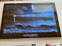 Full view of framed lighthouse picture on board mounted on wall, showing waves, lighthouse, and stormy sky.