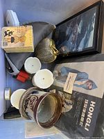 Overall view of box with assorted candles, ceramic pot, brass decorative jar, framed artwork, booklets, and yellow cartoon box.