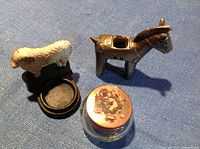 Photo showing a metal sheep candle holder, metal donkey candle holder, and small glass jar with floral lid arranged on a blue fabric surface.