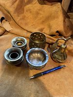 Five small decorative items made of brass and silver placed on a surface with a pen for scale