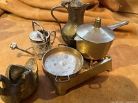 Photo showing miniature brass kitchenware items placed on a brown cloth including a teapot, covered pot, two-handled pan, watering can, and duck creamer.