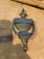 Front view of the brass door knocker showing urn shape, handle, and lettering 'ANAMAKER'