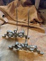 Two brass tripod picture holders placed on a fabric background, showing side view with prongs and ornate bow and ribbon design.
