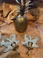 View of complete lot showing brass pineapple container with removable top and two pineapple wall candle holders placed on leather background.
