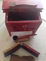 Red wooden shoe shine box with 'Shoe Shine' painted on the front, lid open with shoe cleaning brushes and tools placed next to it.
