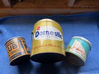 Three antique tin cans including Domestic, Ruby Tobacco, and Player's brands without lids or holes placed on a blue surface.