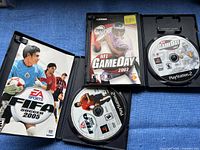 Two PlayStation 2 game cases opened and laid flat showing discs inside: FIFA Soccer 2005 and NFL GameDay 2002.