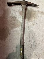Full view of vintage pick mattock tool standing on concrete floor, showing wooden handle and metal pick head.