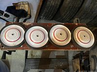 Four metal wagon wheels placed in a row on a wooden surface, showing front face with off-white paint and red concentric rings. Background includes tires and workshop area.