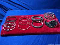 Image of 11 mixed silver-tone bracelets arranged on a red velvet display, showing variety in design including thin bangles, filigree cuff, and colored stone cuff.