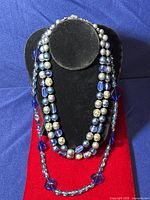 Two blue glass necklaces displayed on black and red stands against a blue background. One necklace is multi-strand with mixed beads, the other is single-strand with faceted blue beads.