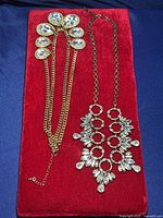 Two gold tone crystal necklaces displayed on red cloth and blue background.