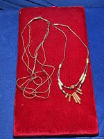 Two Indigenous American liquid silver necklaces placed on a red velvet surface; one has dangling goldstone pendants and shell beads, the other is a plain silver chain tangled and in need of repair.
