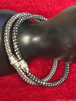 Two tubular bracelets adorned with small metallic stones, one darker and one lighter silver tone, shown coiled on a black statuette against a red background.