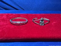 Two sterling silver bracelets displayed on red velvet background against blue fabric. One is a solid bangle with geometric cutout, the other an open cuff with seated figure.
