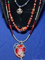 Three Indigenous American style necklaces displayed on a black velvet necklace bust against blue background. Visible necklace details include a prominent red glass wire-wrapped pendant, red wood and resin beads, and a smaller silver pendant with blue glass beads.