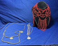 The three necklaces displayed with the red beaded one on a black display bust for presentation, two silver-tone coin pendant necklaces laid beside on blue fabric background.