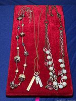Three mixed random necklaces laid on a red background: a silver-tone decorative bead necklace, a gold-tone ball chain with pendants including bars and a heart, and a chain with large clear and white beads.
