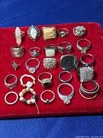 Rings arranged on red velvet surface showing varied styles and materials including silver and gold tones, stones and textured bands.
