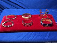 Photo of six different gold tone bracelets arranged on red fabric showing variety in design and size.