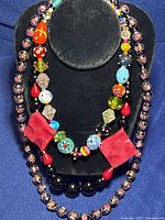 Five bead jewelry items displayed on black stand: two Murano glass bead necklaces, one black onyx bead necklace, one red agate necklace, one red coral and glass bead bracelet.