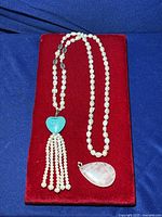 Full necklace displayed laid on red velvet showing pearl strand, turquoise heart, and pearl tassels with rose quartz pendant next to it.