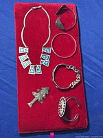 Photo showing alpaca turquoise necklace, four silver plated bracelets, and mixed metals Christmas tree brooch laid on red velvet fabric.