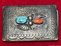 Close-up front view of rectangular sterling silver belt buckle with hammered texture, etched border, one turquoise stone, one coral stone, silver fan shapes and beads.