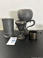 Group photo showing four antique metal pieces: large three-handled loving cup, smaller three-handled loving cup, mid-19th century pewter tankard, and small silverplate cup.