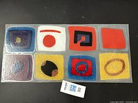 Clear rectangular glass panel with nine colorful abstract geometric shapes arranged in a 3x3 grid, photographed flat on a black background.