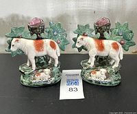 Pair of 19th century Staffordshire spill vases featuring standing cows with red-brown patches and recumbent calves, surrounded by green foliage with floral accents and tree trunk style spill holders with pink interiors, placed on black surface with auction lot number.