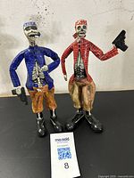 Front view of two hand-painted papier-mâché skeleton figurines, one in blue and orange, the other in red and brown, showing detailed skeletal torsos and expressive faces.