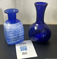 Both glass vessels displayed side by side on a plain surface including the textured frosted cobalt glass bottle with etched wave motif and the cobalt blue handblown vase.